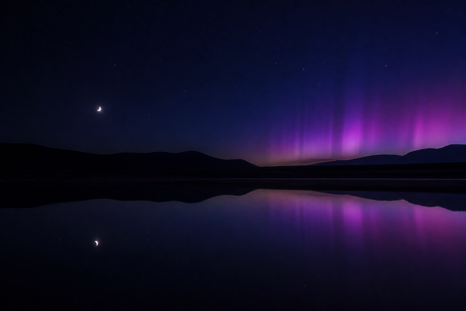 A crescent moon over a still lake, subtle violet aurora on the horizon